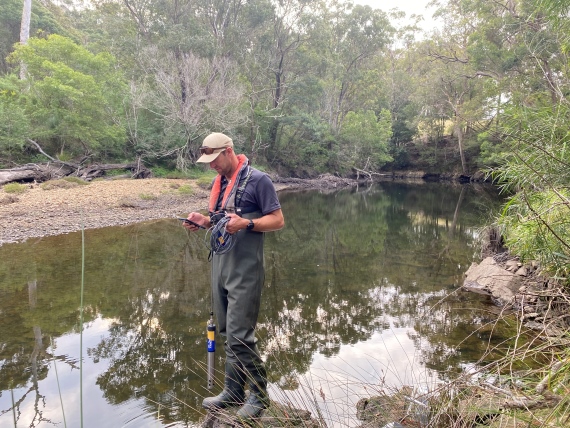 A researcher stands on the edge of an estuarine creek testing salinity levels using a hand held probe.
